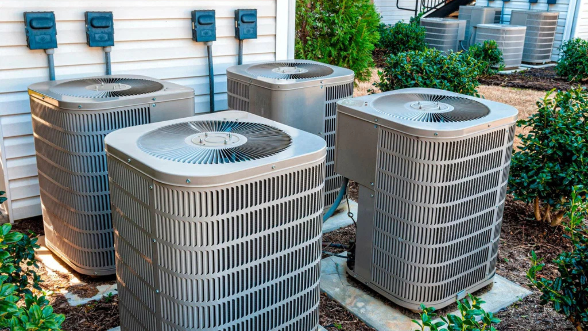 Four outdoor air conditioning units in a yard next to a building.