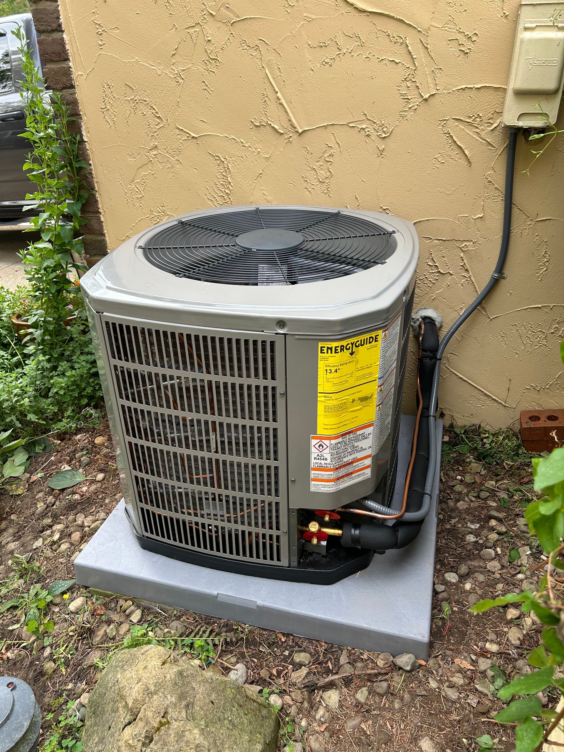 Air conditioning unit outdoors on concrete base, next to a stucco wall and greenery.