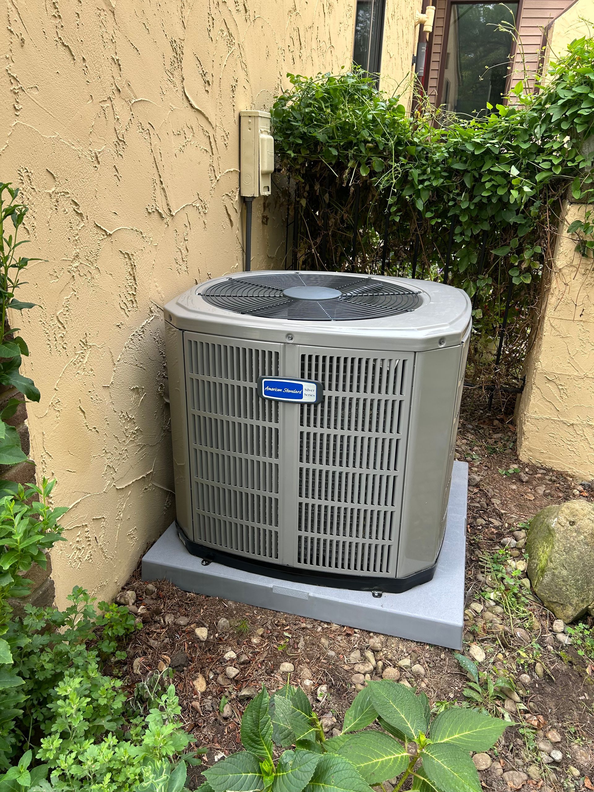 Air conditioning unit on a concrete pad, next to a stucco wall and bushes.