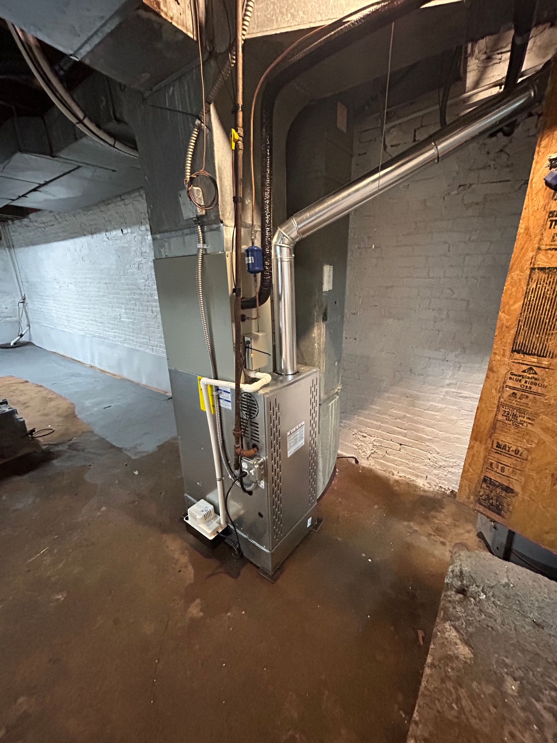 Furnace in a basement. Ductwork and pipes are visible. Floor is concrete and wet.