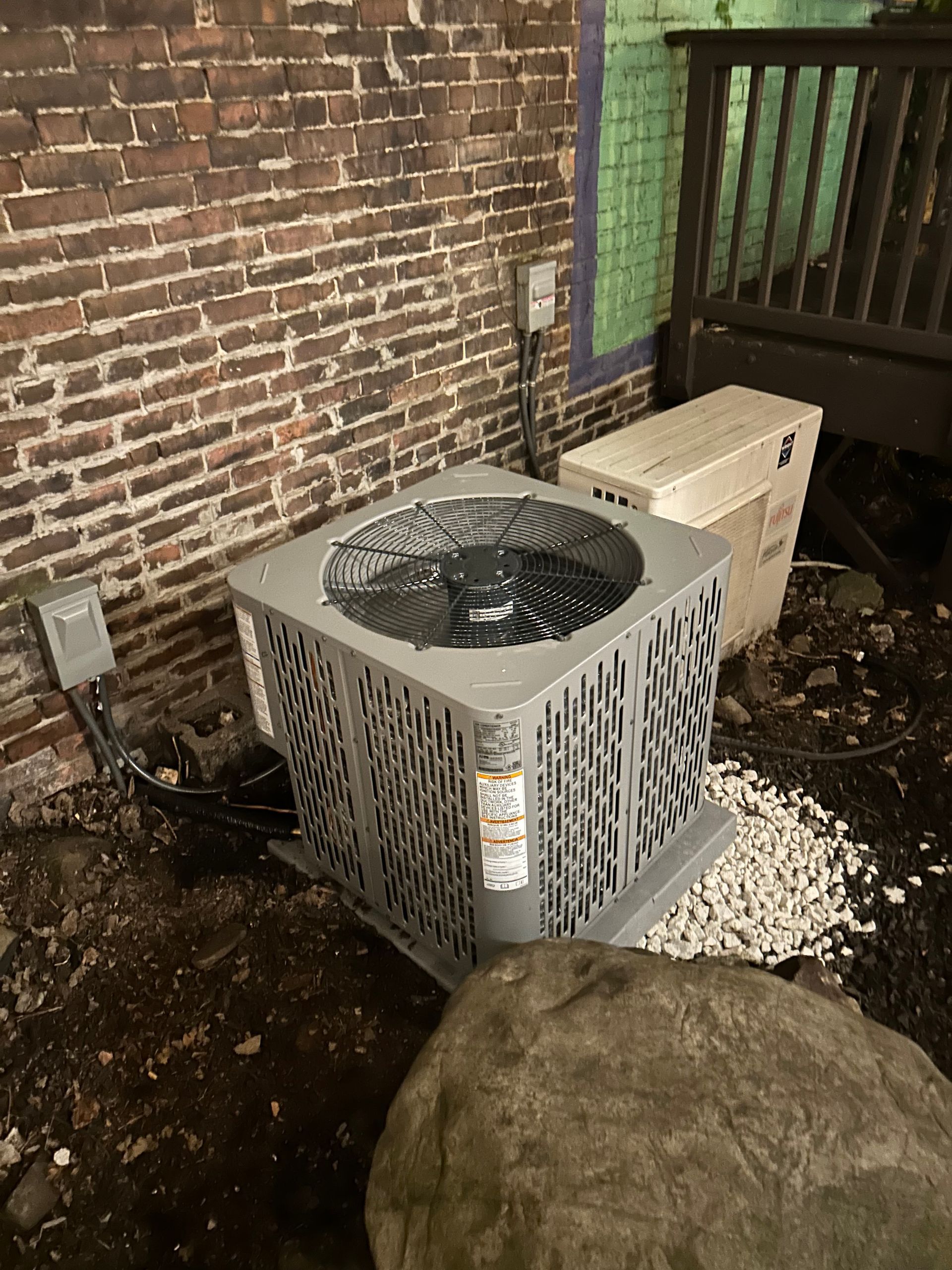 Air conditioner unit on gravel near a brick wall and a wooden railing.