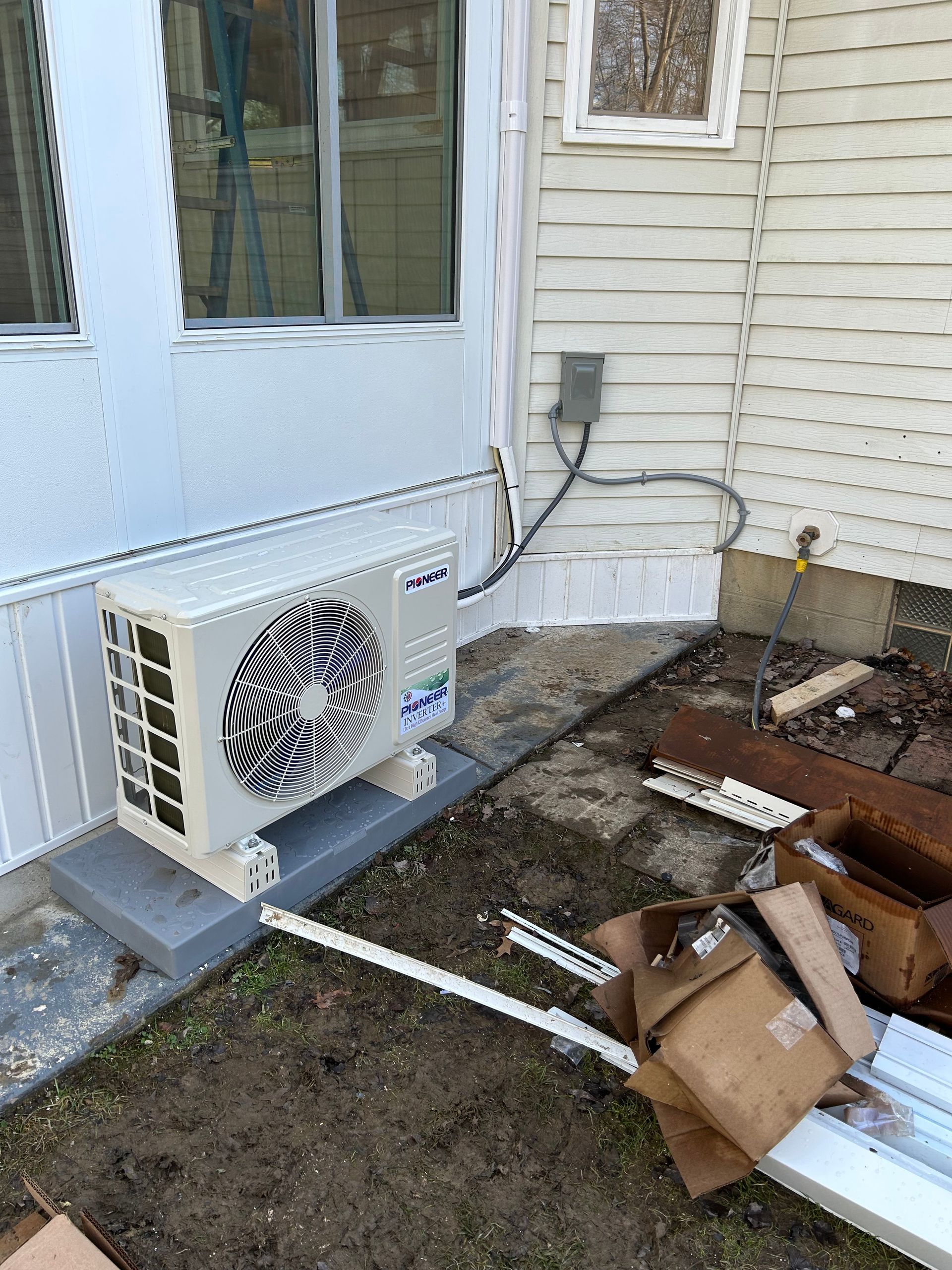 An air conditioning unit sits on a concrete pad next to a house. Debris is scattered around it.