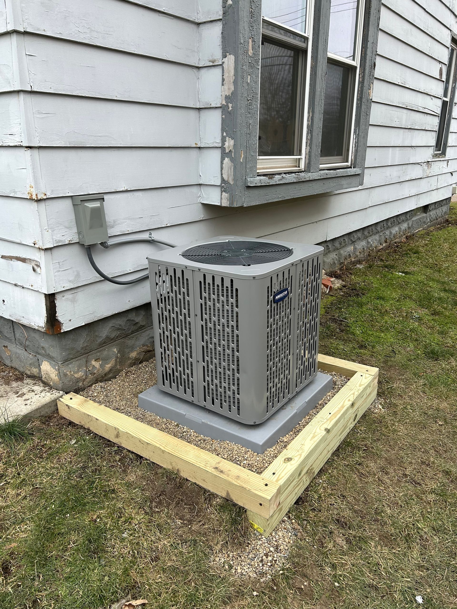Gray AC unit on a concrete pad next to a house with a wooden frame base.