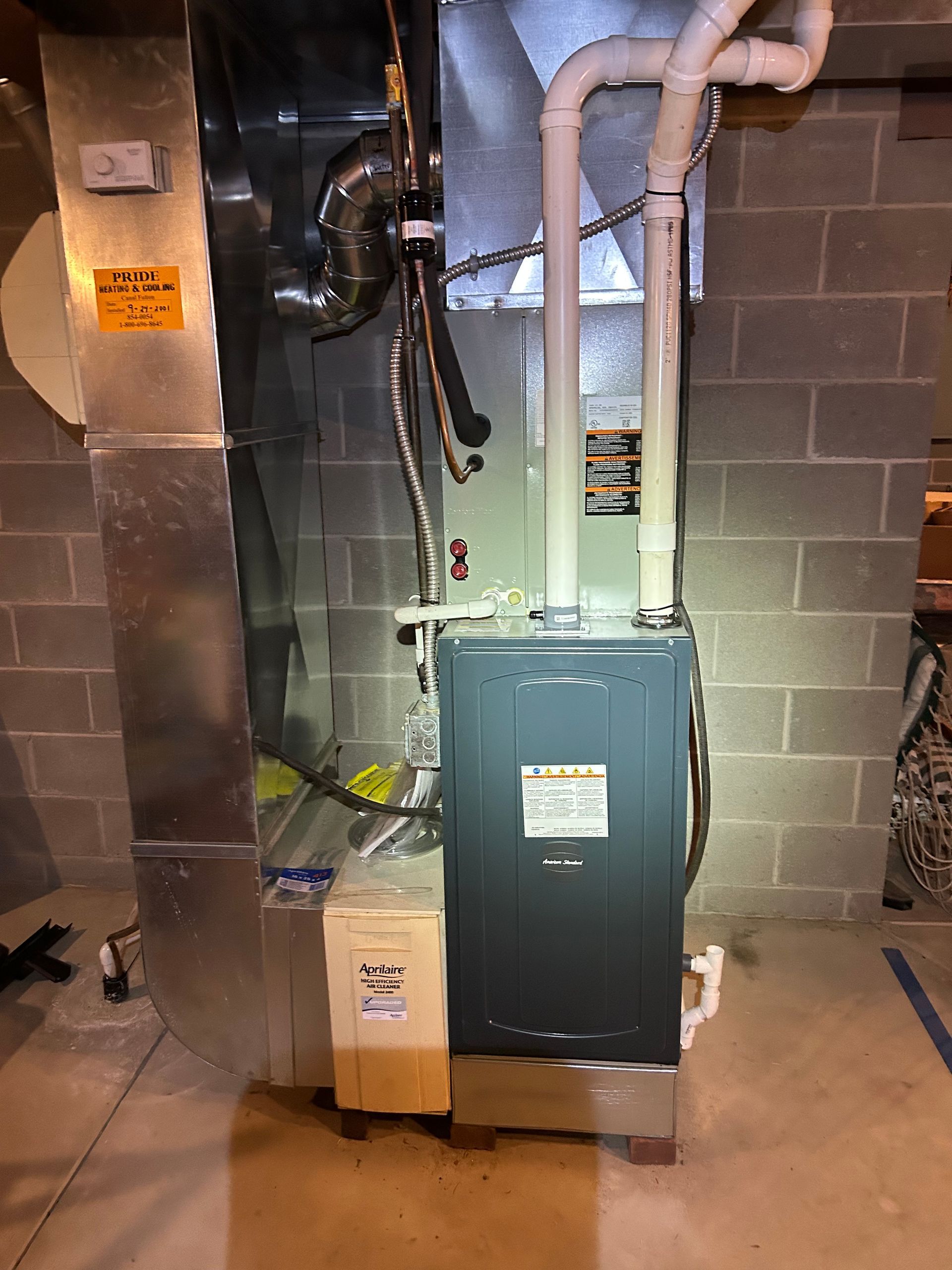 A furnace in a basement setting. It is gray and tan, with ductwork and ventilation pipes attached.