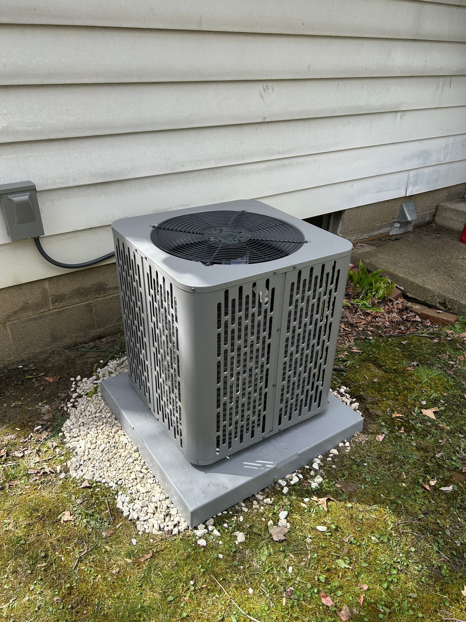 Gray air conditioning unit on a concrete pad, next to a white siding wall and grass.