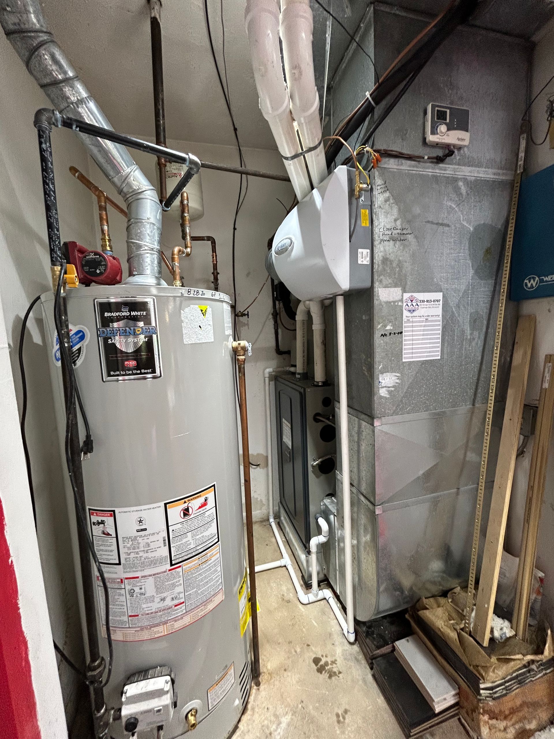 Water heater and furnace in a basement. Grey metal and pipes, with a white humidifier on top.