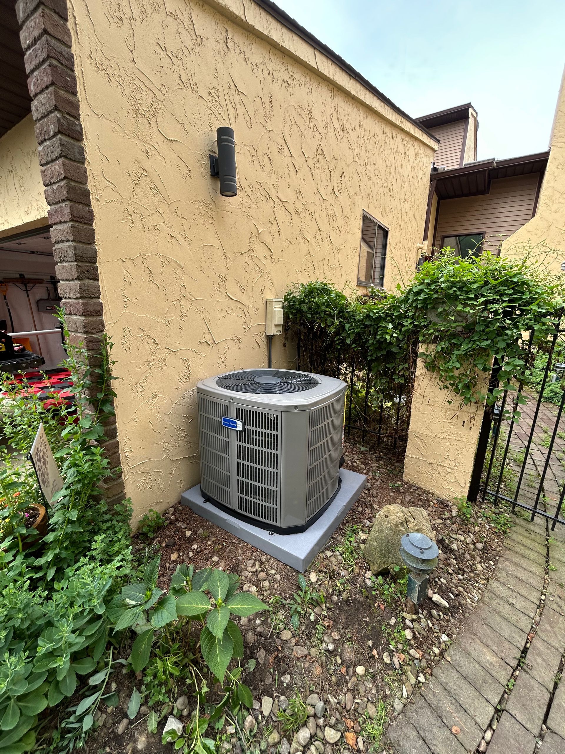 AC unit on a concrete pad, surrounded by greenery, next to a tan stucco building with a brick column.