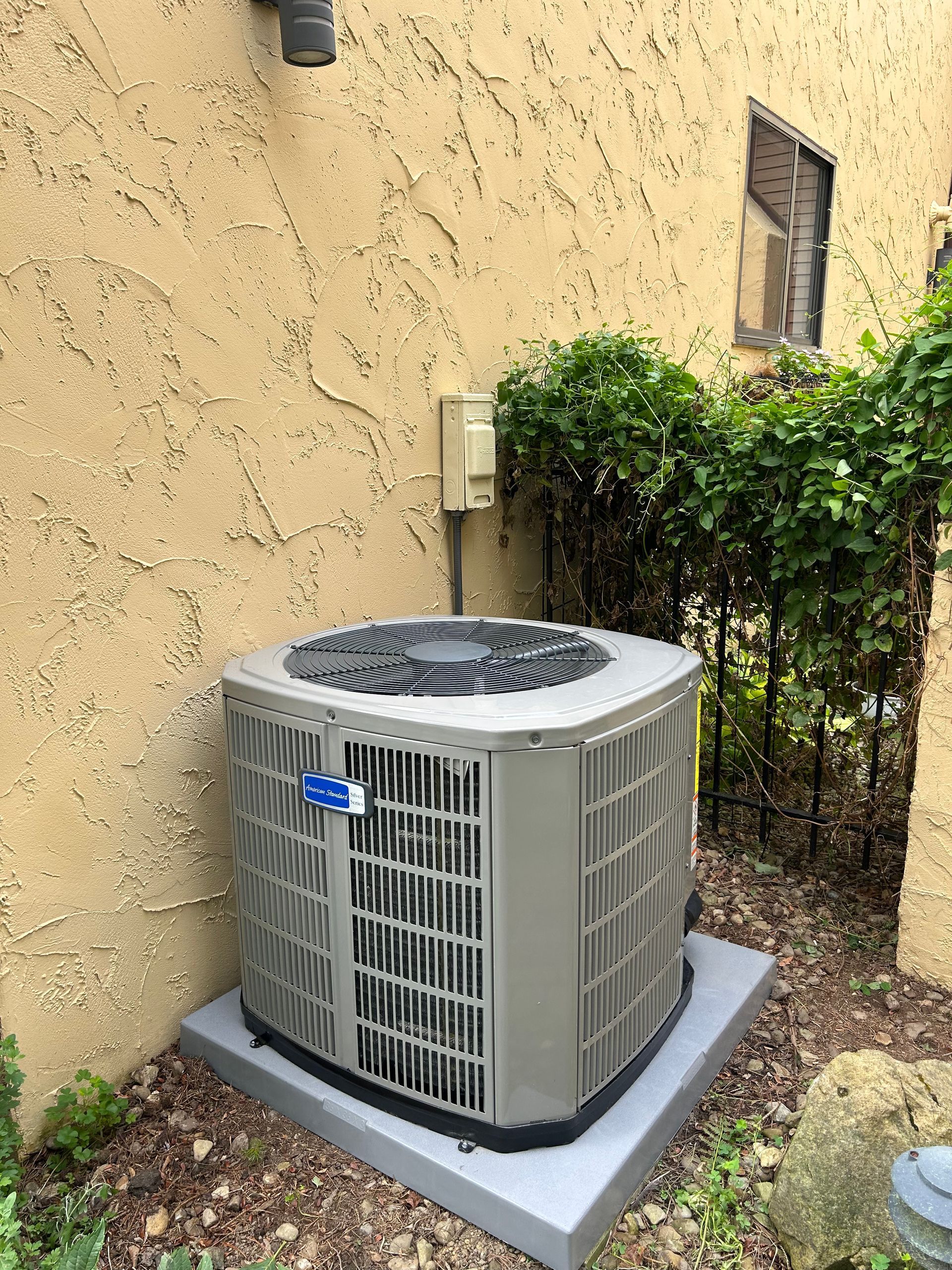 An air conditioning unit outside a tan stucco wall. Green bushes and a small window are behind it.