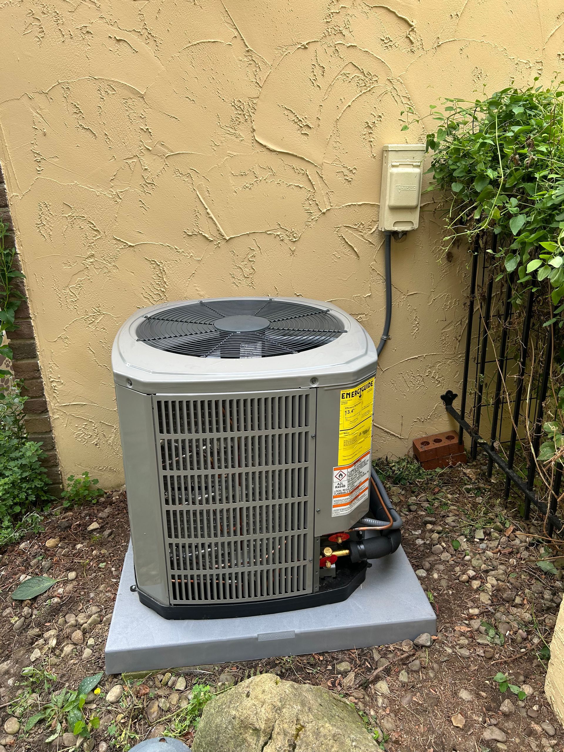Air conditioner unit on a concrete pad next to a stucco wall and shrubbery.