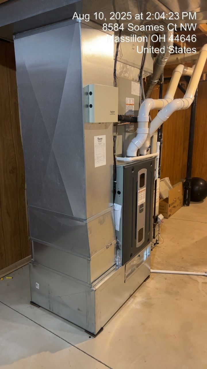 A tall, silver HVAC unit in a basement, with white pipes and a visible filter.