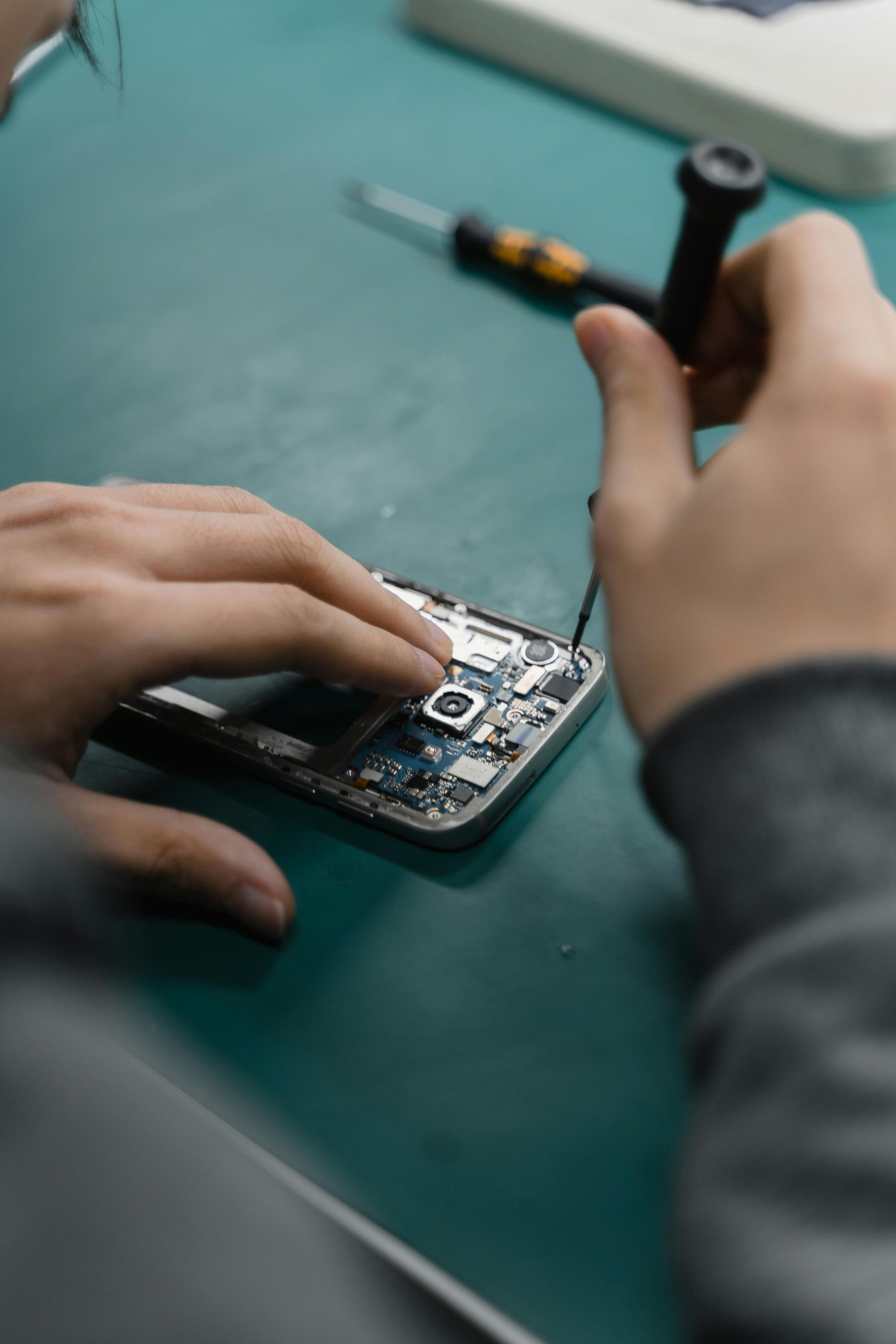 Person repairing a mobile phone with a screwdriver on a green surface.