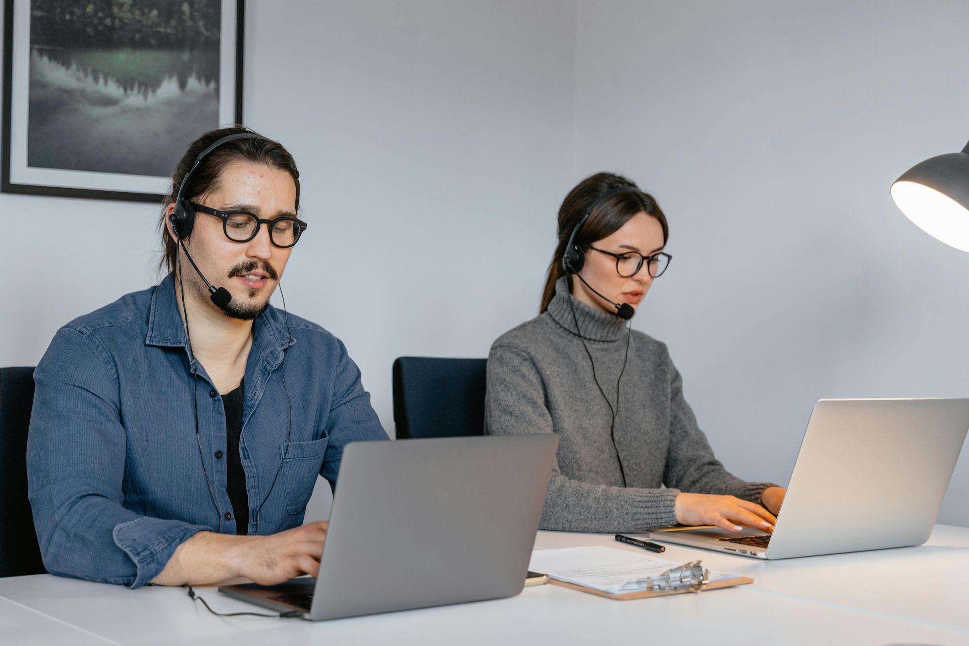 Two people wearing headsets, working on laptops at a desk.