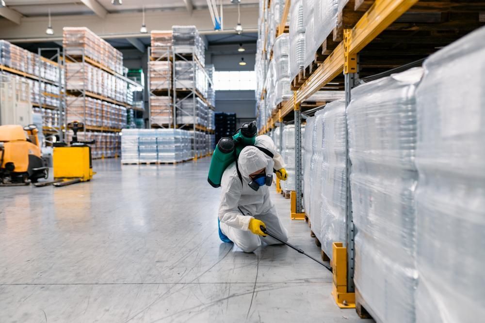 A Man In A Protective Suit Is Spraying A Warehouse With A Sprayer — X-Terminate Pest Control In Imbil, QLD