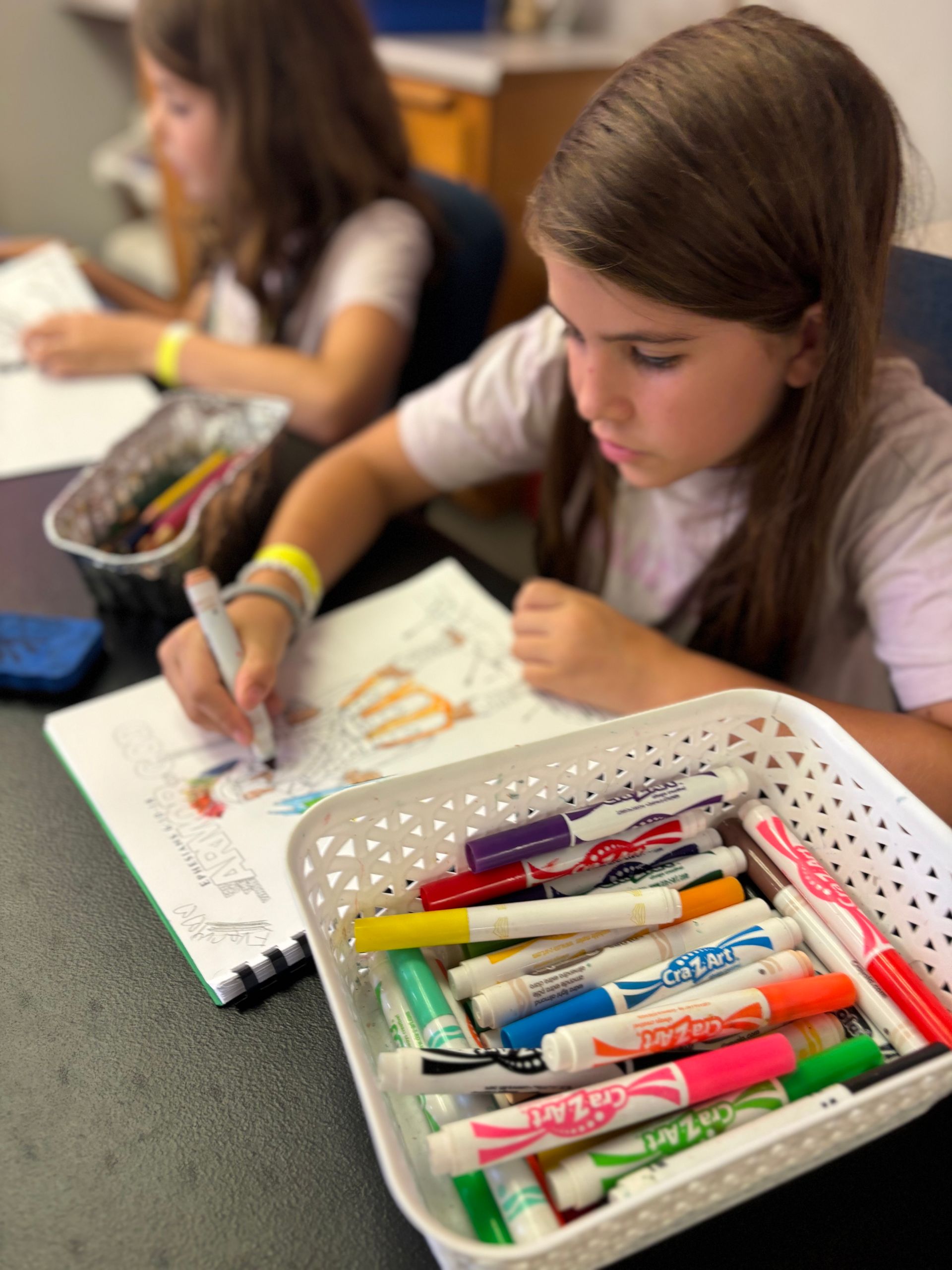 A girl is sitting at a desk with a basket of markers in front of her