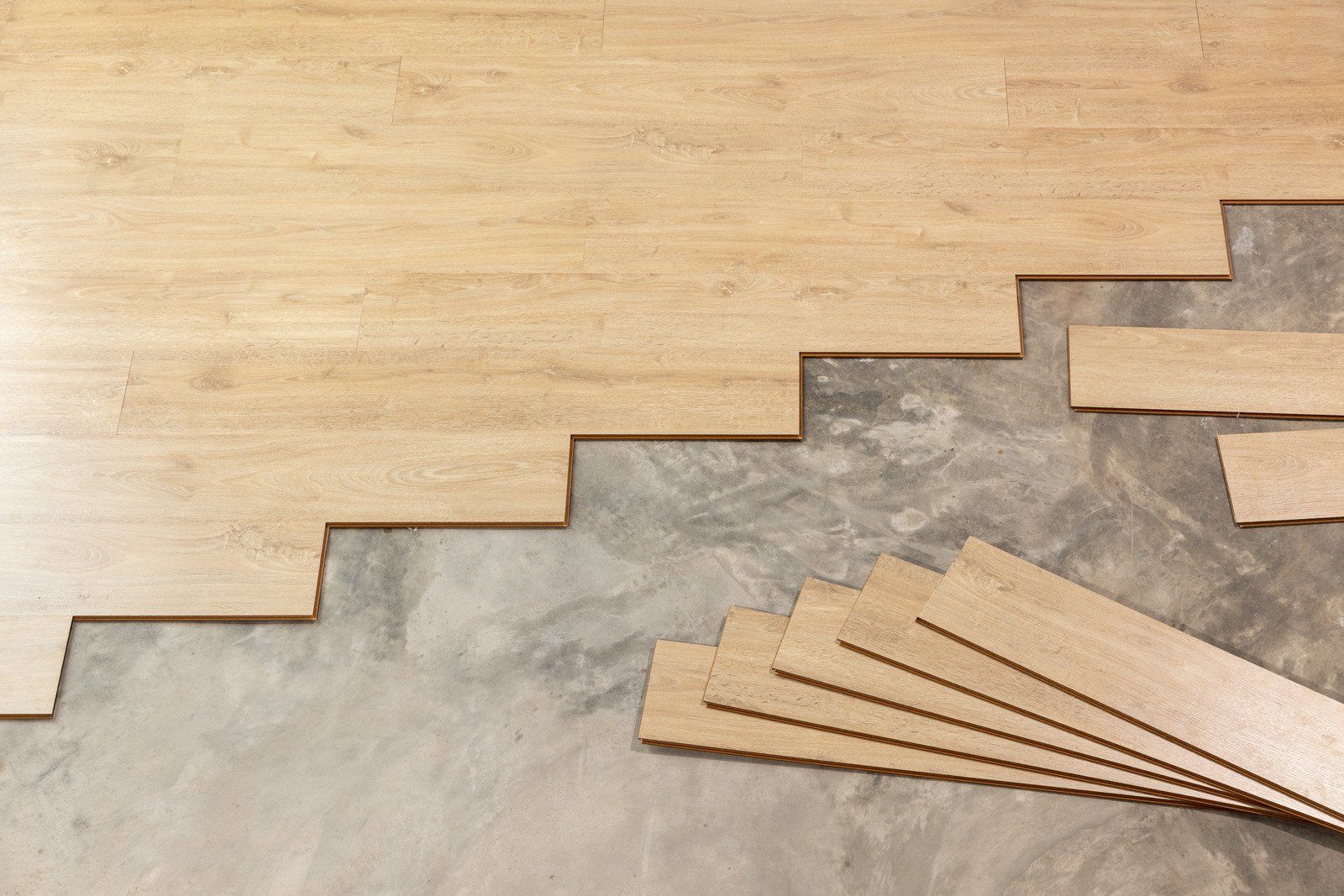 Wood flooring installation in progress on a concrete subfloor; light brown planks partially cover the surface.