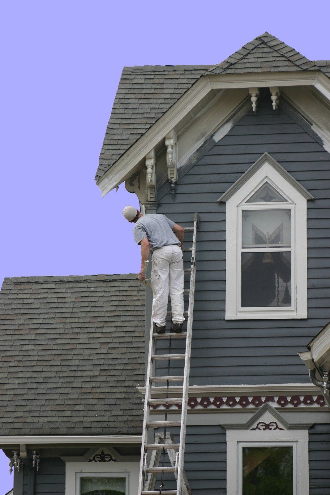 Person on a ladder painting the exterior of a blue Victorian-style house.