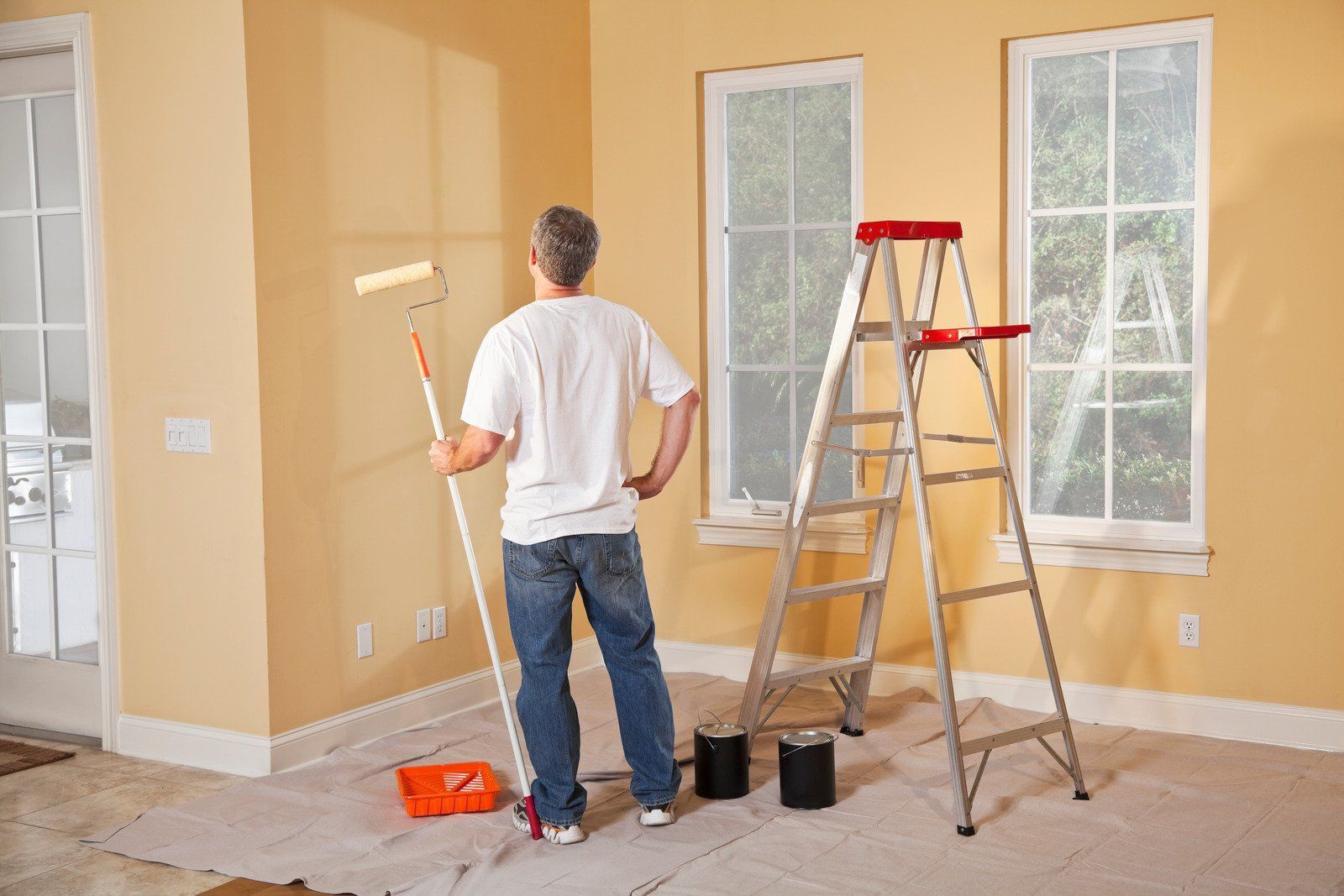 Man painting a room with yellow walls; he stands with a paint roller, near a ladder, paint cans, and an orange tray.