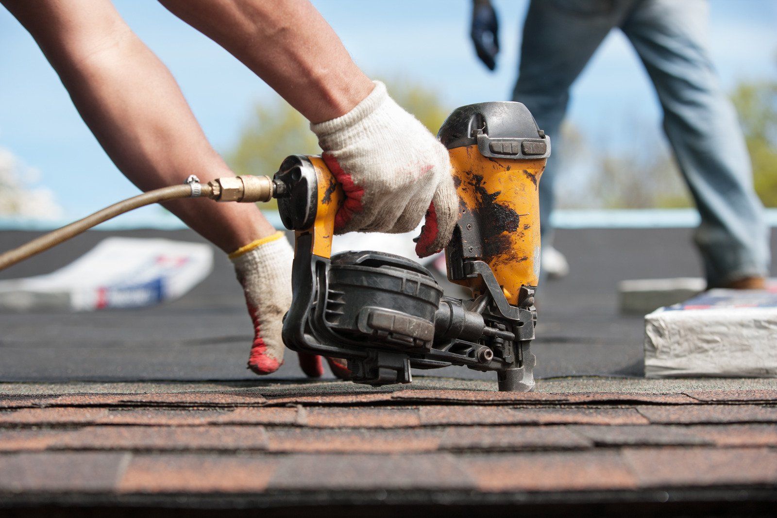 A person using a nail gun on a roof, installing shingles. The person wears gloves; a second person is in the background.
