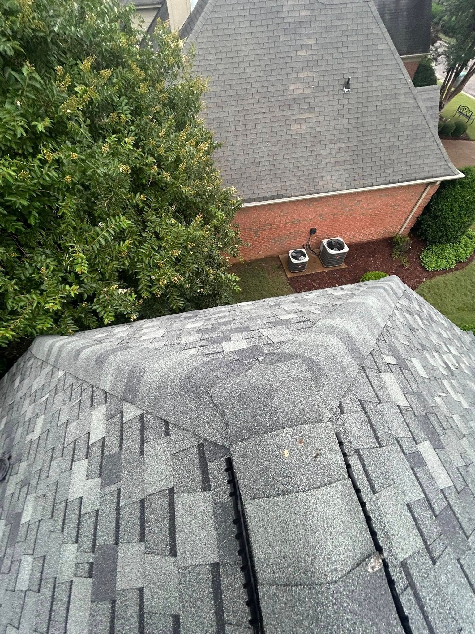 Overhead view of a house roof with dark gray shingles, next to a tree and a brick wall.