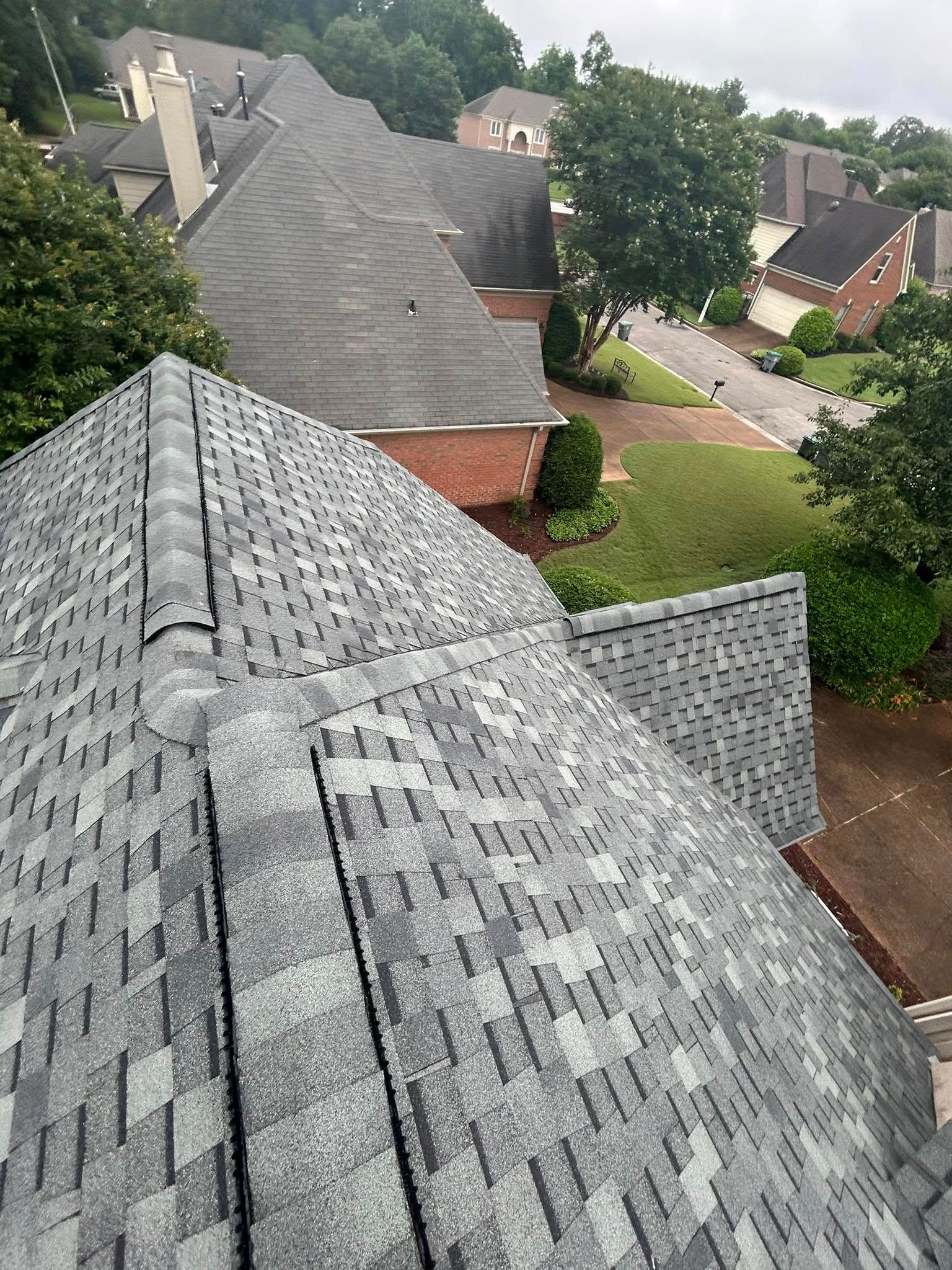 Overhead view of a gray shingled roof with surrounding houses, greenery, and a street visible in the distance.