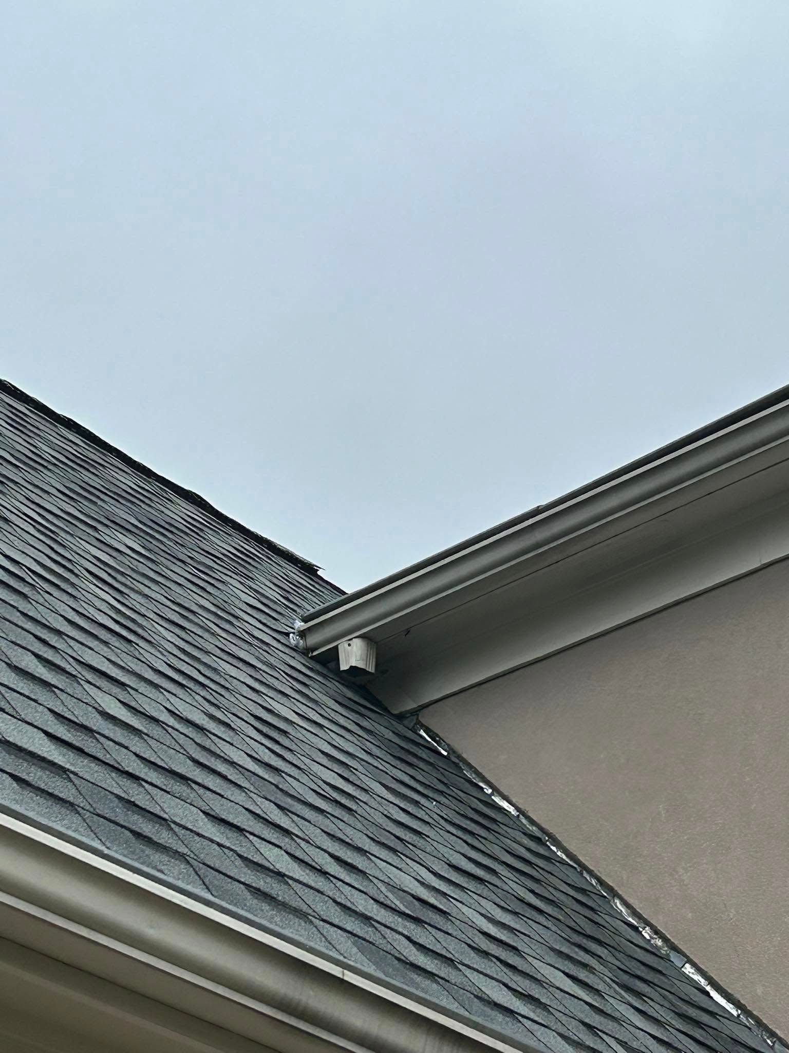 Gray-shingled roof and beige siding meeting. A security camera is mounted on the corner near the roofline. Overcast sky.