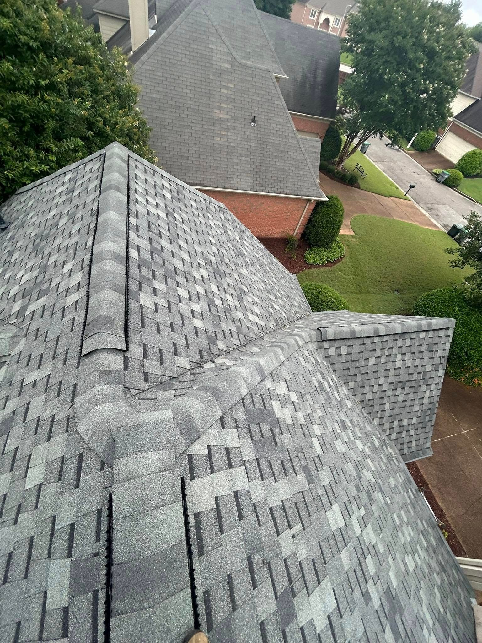 Overhead view of a gray shingled roof on a house. Green foliage and other rooftops are visible in the background.