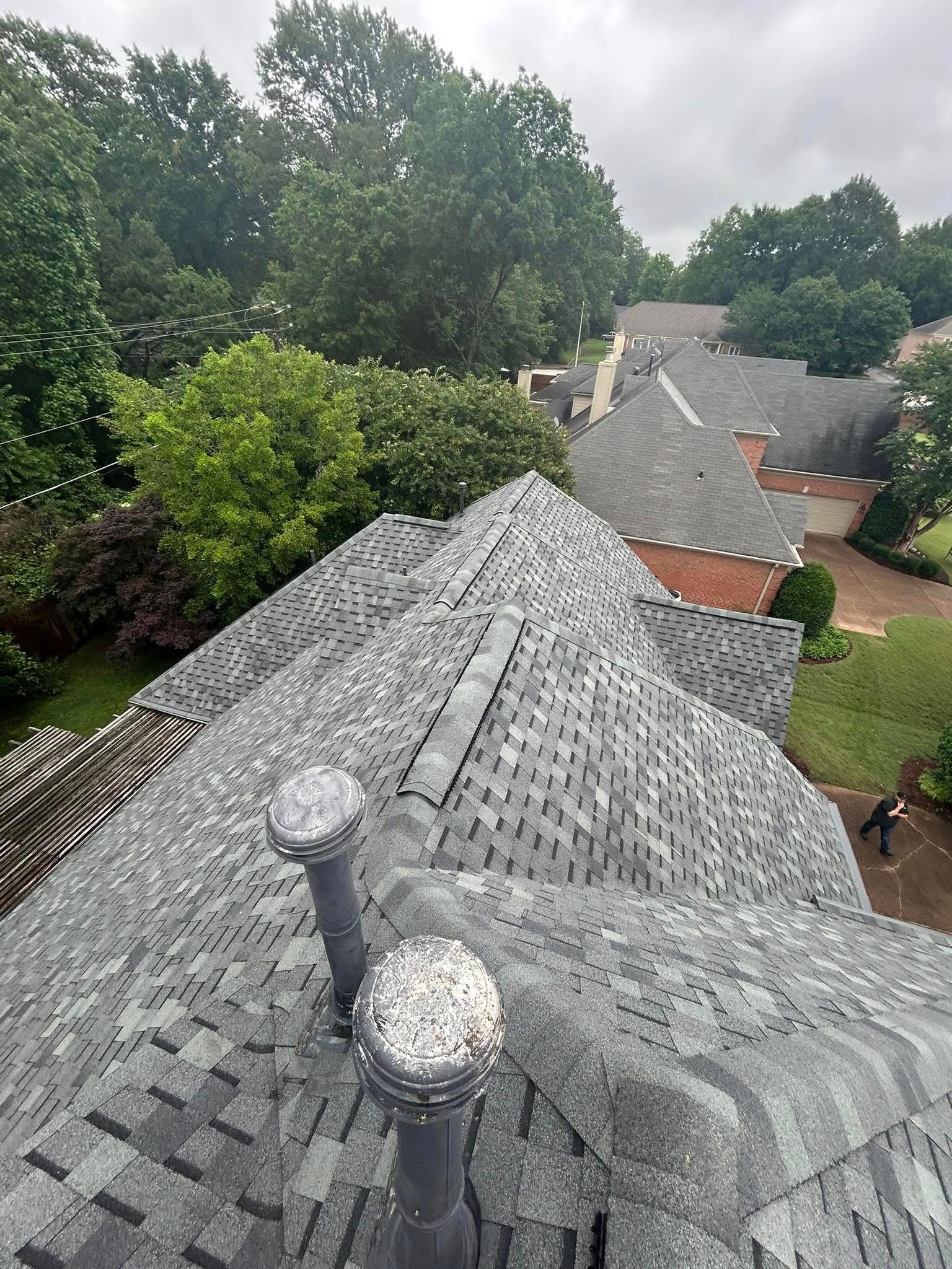 Overhead view of a gray shingle roof with two chimneys, lush trees in the background, and neighboring houses.