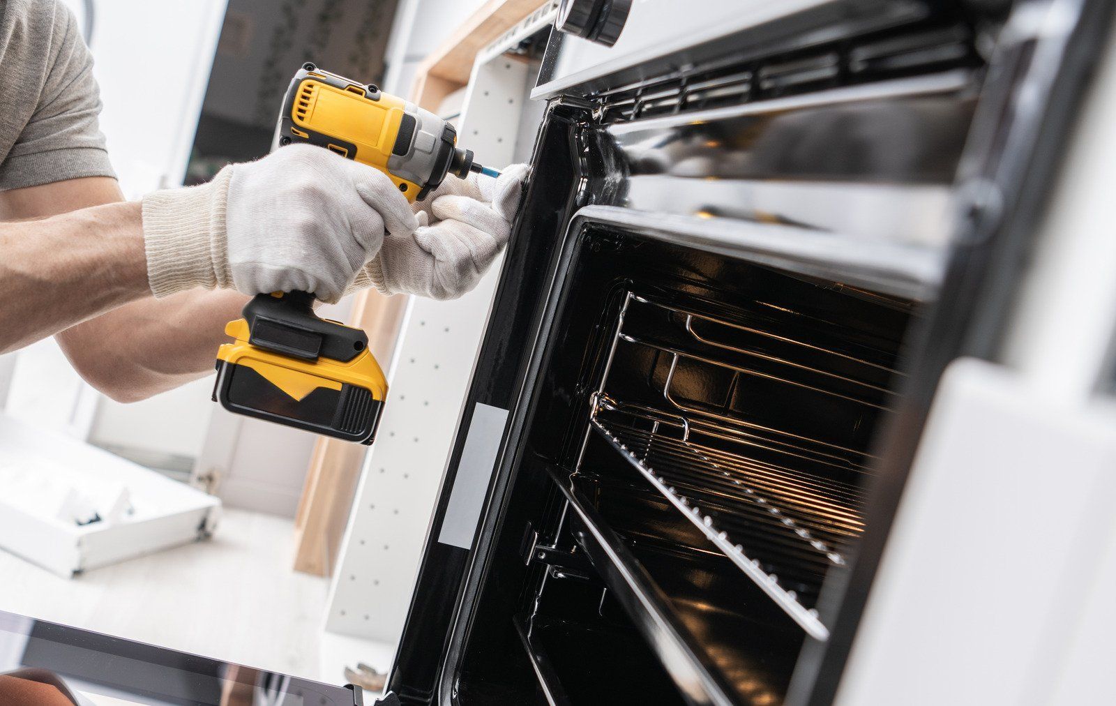 Person in gloves using a yellow power drill to work on an oven in a kitchen.