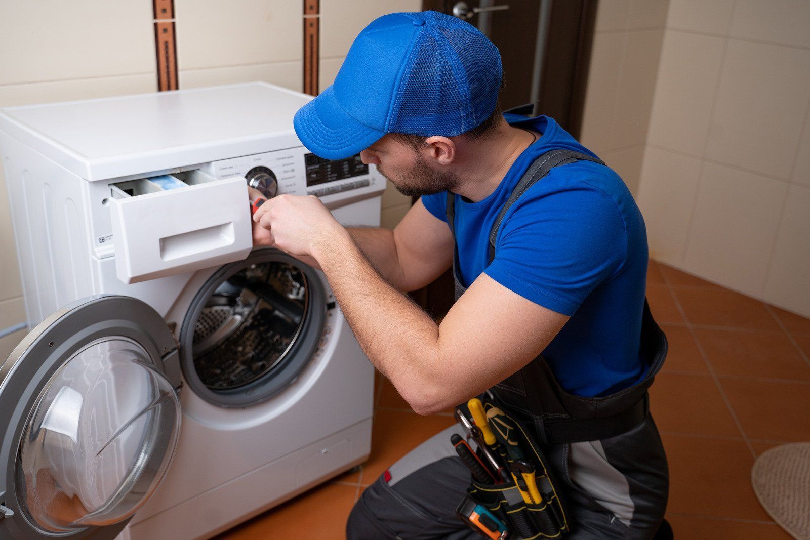 A repairman in blue overalls and a cap repairs a white washing machine in a tiled bathroom.