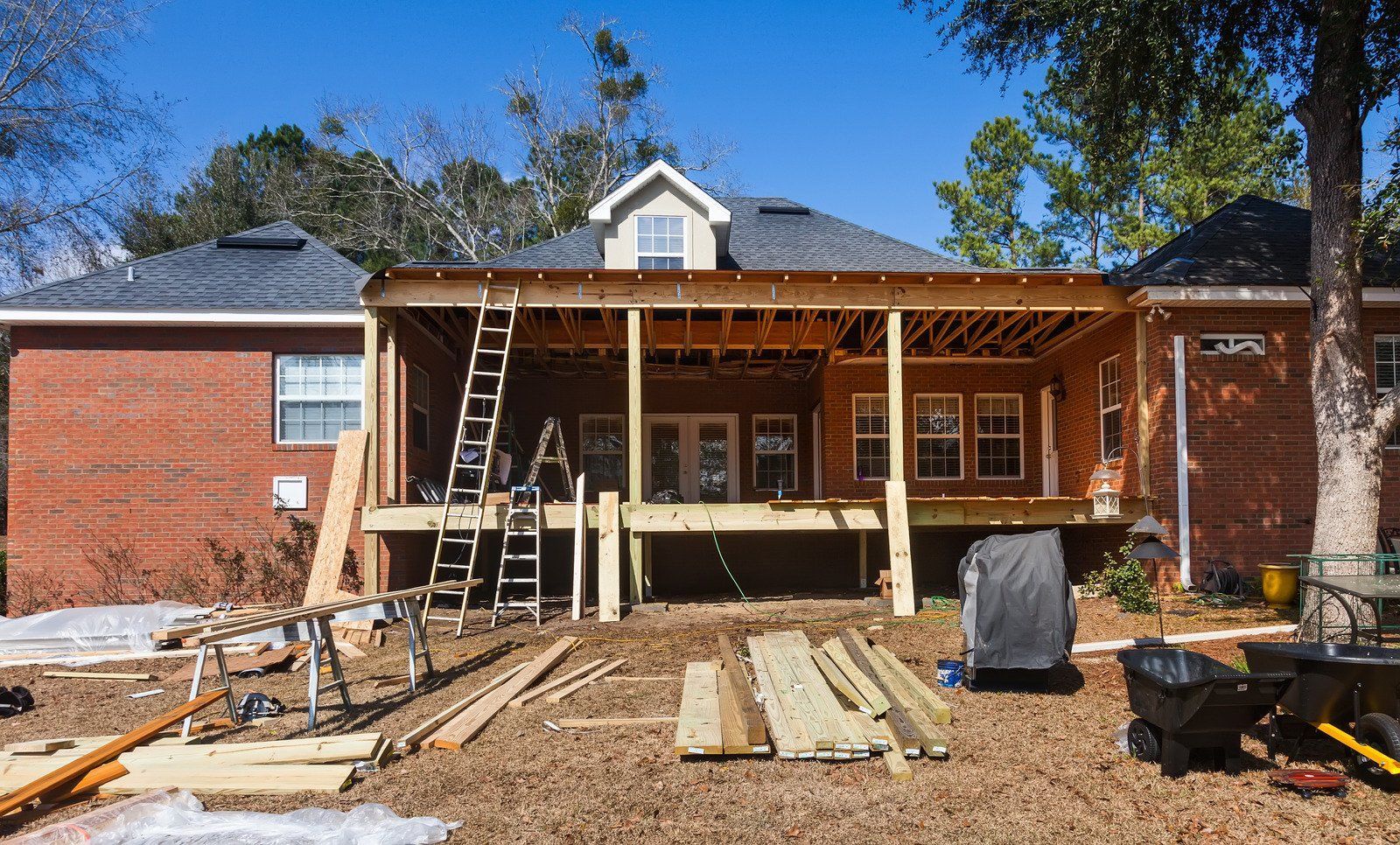 Backyard construction of a deck attached to a brick house. Lumber, ladders, and tools scattered around the yard.