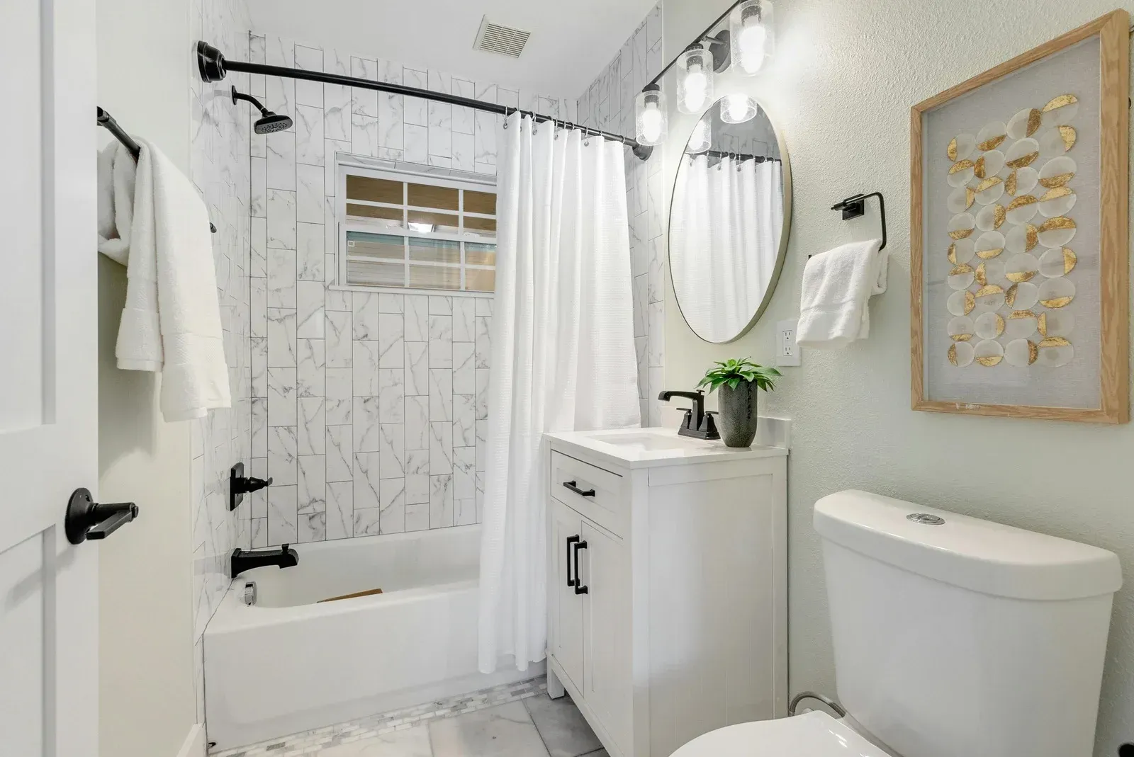 White bathroom with shower, vanity, and toilet. Features marble tile, black fixtures, and a framed artwork.
