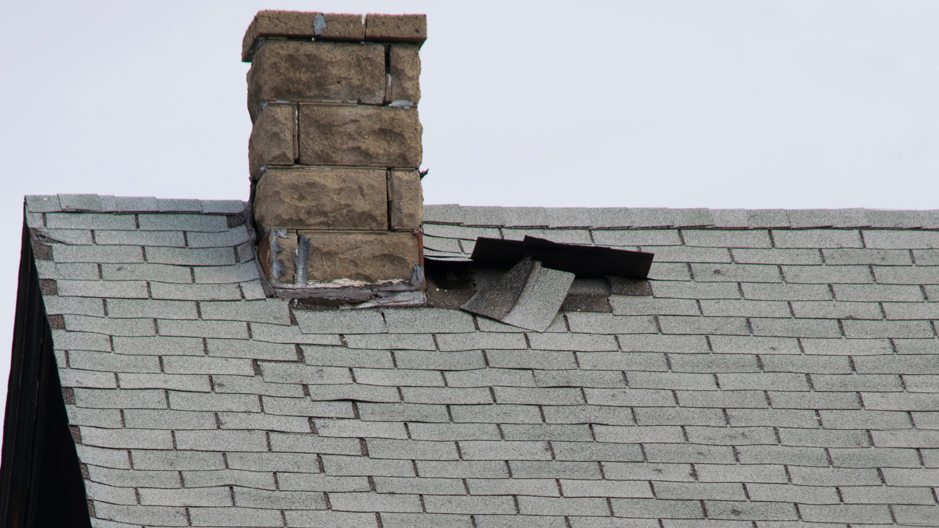 Damaged asphalt shingle roof around a stone chimney, gray sky background.