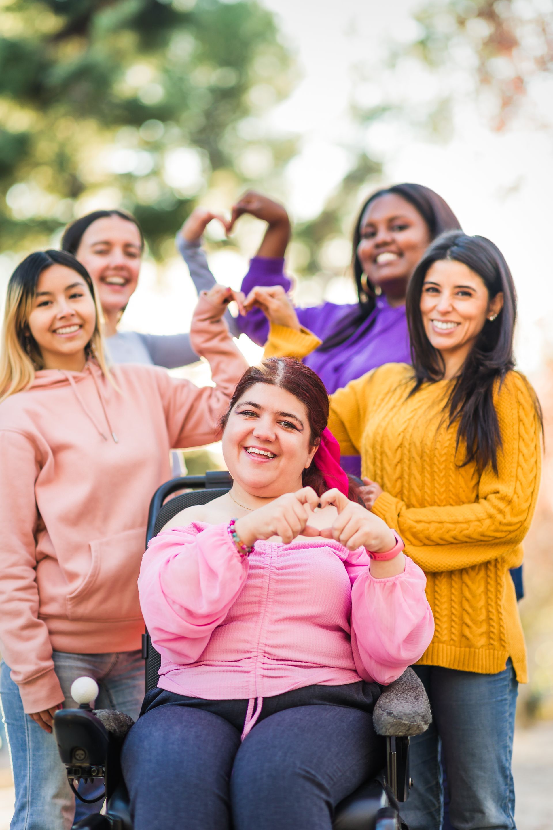 A group of women are standing around a woman in a wheelchair making a heart shape with their hands.