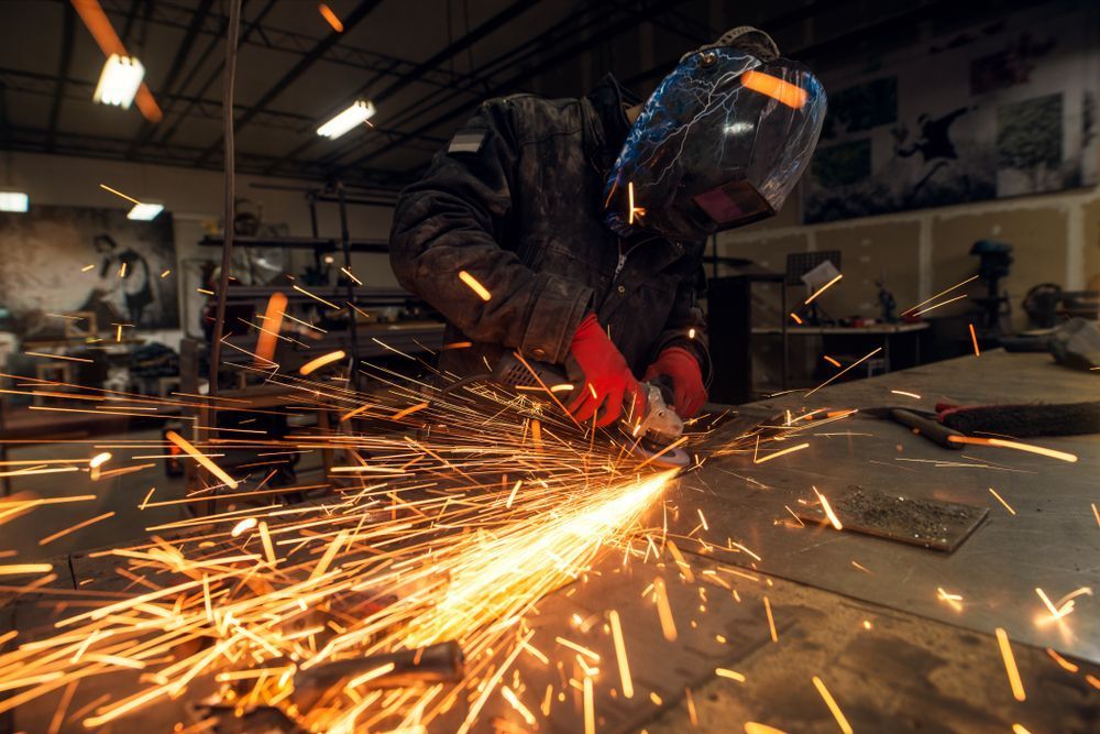 Welder In A Workshop, Sparks Flying From Metal Being Ground. Red Gloves, Protective Helmet