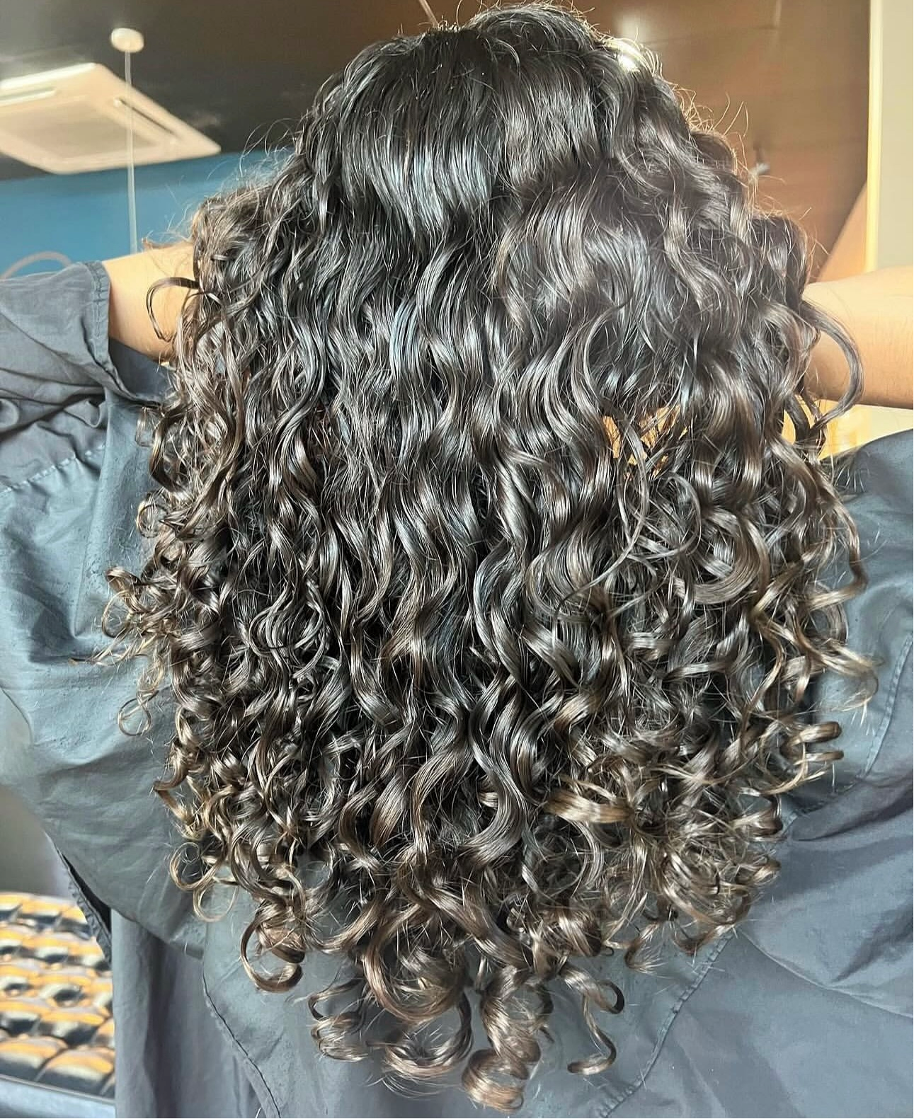 Woman with shiny, dark, curly hair. Back view, salon setting.— Gina T Hair & Beauty In Mackay, QLD