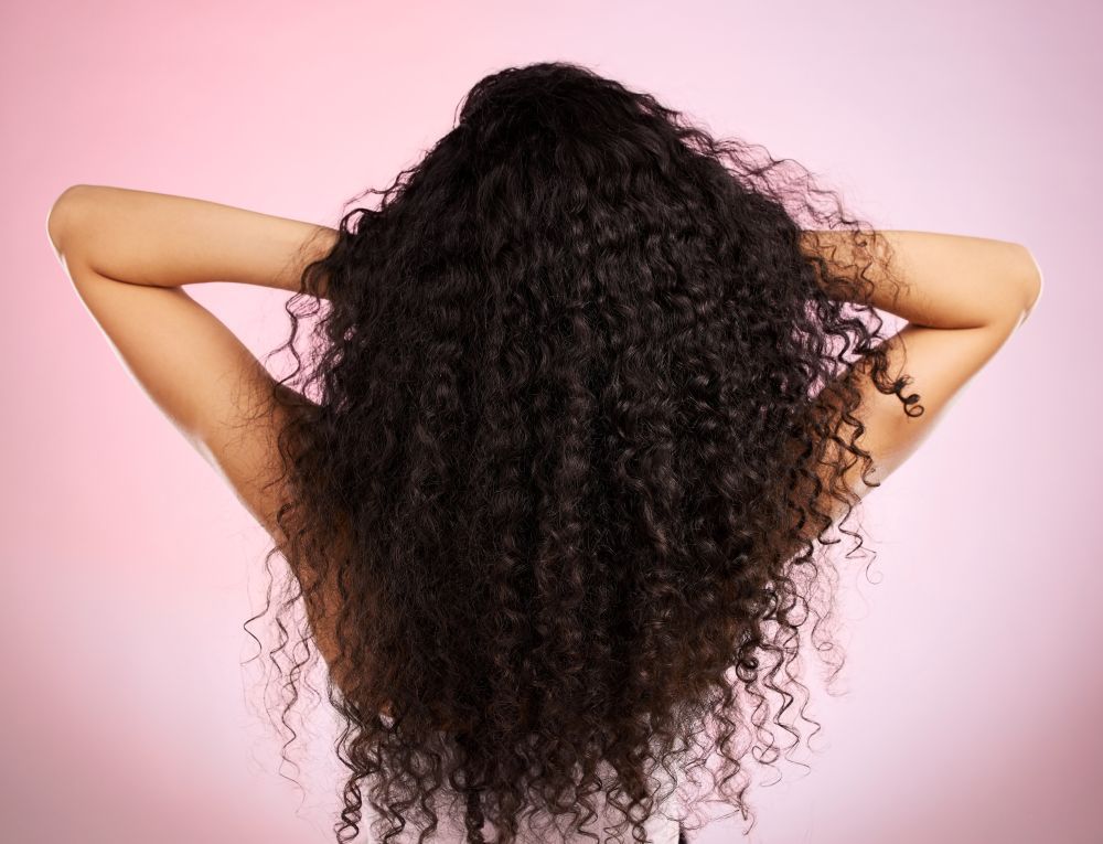 Woman With Dark Curly Hair, Arms Raised, Against a Pink Backdrop — Gina T Hair & Beauty In Mackay, QLD