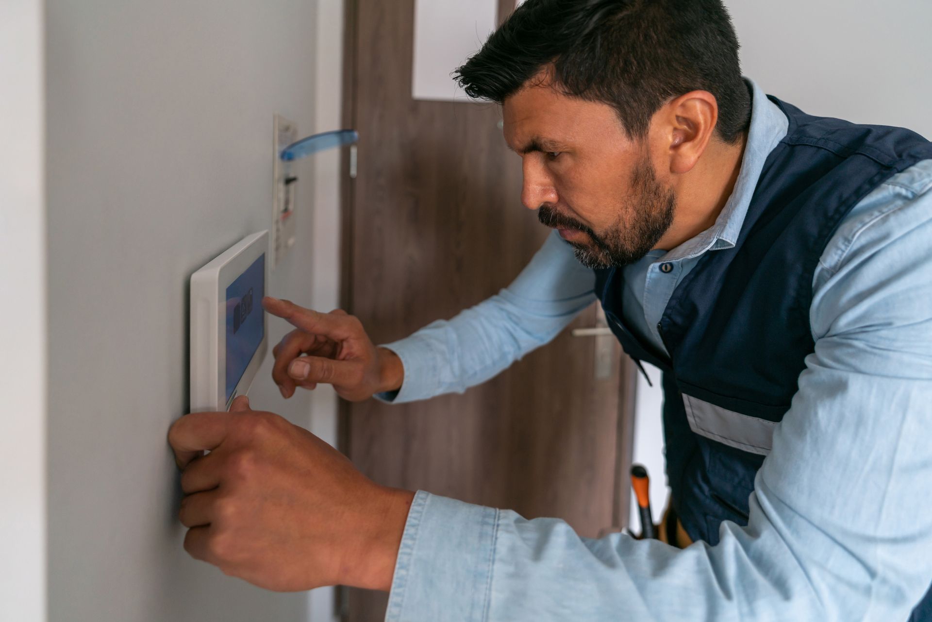 A male technician wearing a blue shirt is installing an alarm system in a residential entrance.