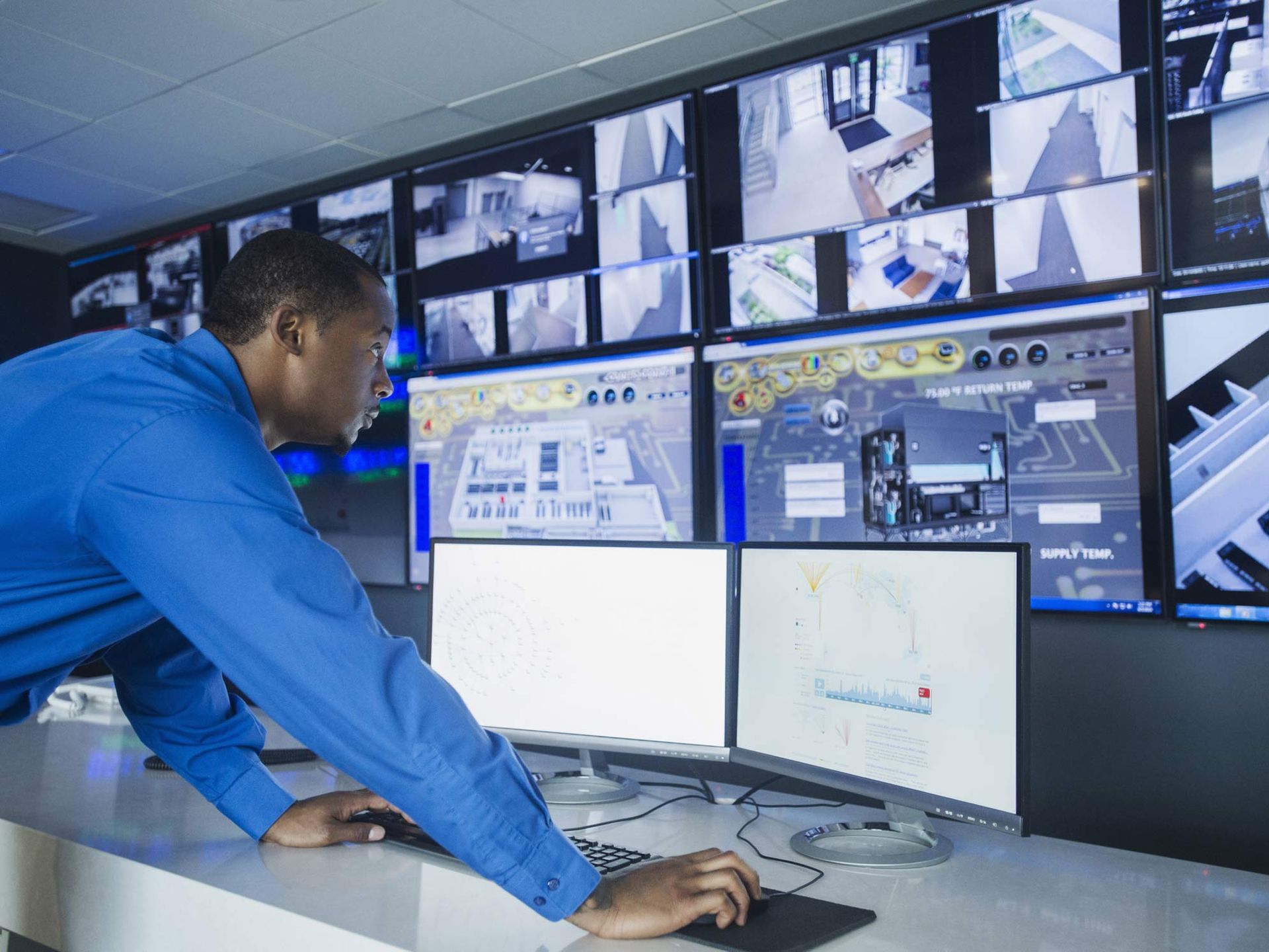 Technician in blue shirt working at a high-tech control room desk.