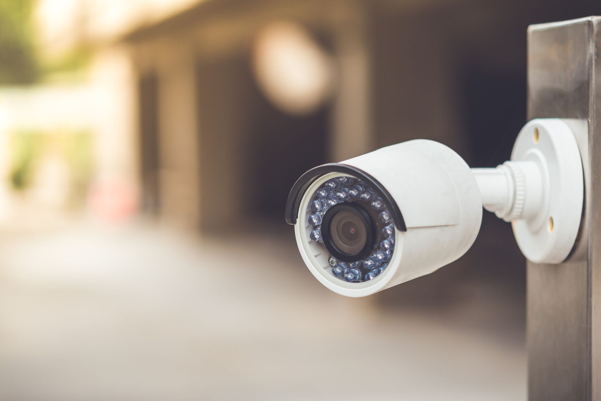 White security camera, mounted on a metal pole, overlooking a pedestrian area with blurred background.