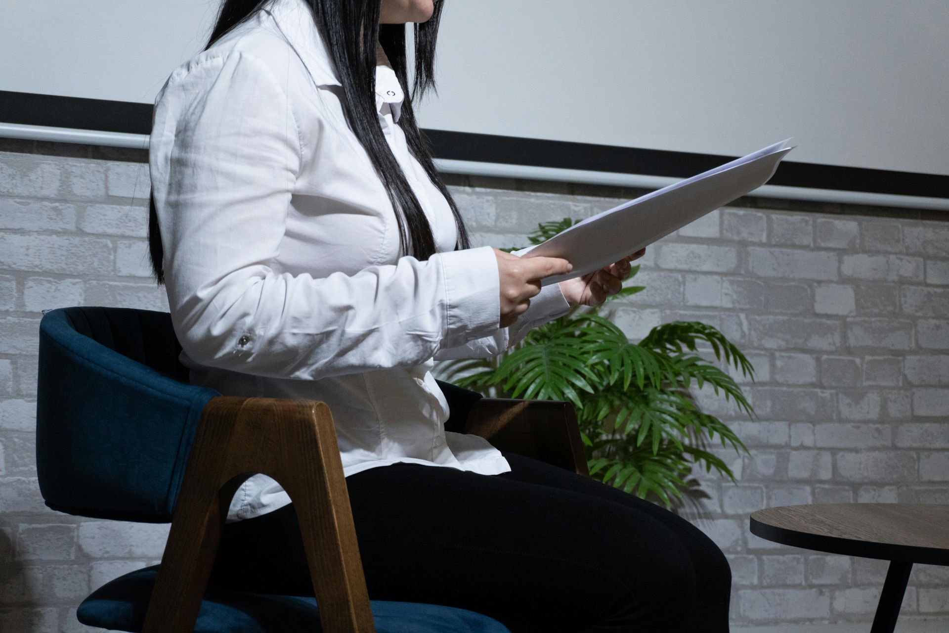 Mulher de camisa branca e calça preta sentada, segurando papéis, perto de uma planta e uma parede de tijolos.
