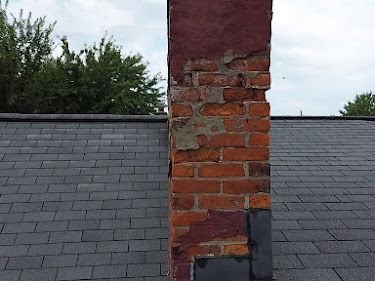 A deteriorating brick chimney on a dark gray shingle roof with visible damage to the mortar and brick..