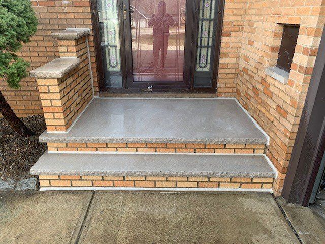 Brick and concrete front porch with two steps leading to a glass door. The porch has a light colored brick and concrete 