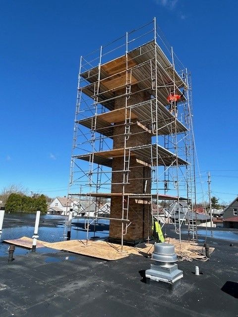 A tall brick chimney surrounded by scaffolding on a flat roof under a bright blue sky.