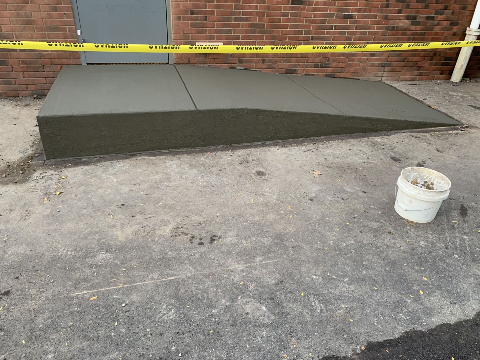 A newly constructed concrete ramp in front of a building with yellow caution tape. A bucket sits nearby.