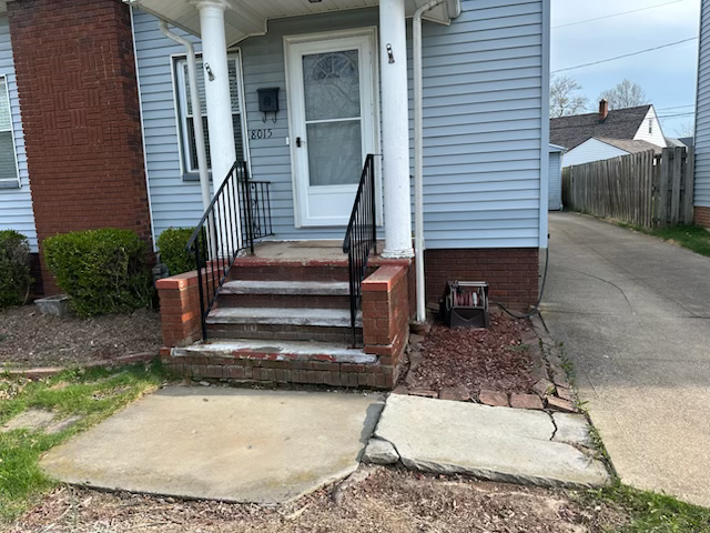 Front entrance of a light blue house with a cracked concrete walkway and steps leading to a front door.