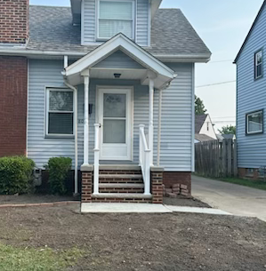 A two-story house with a porch. Gray siding, white trim, and a brick foundation. The yard is freshly tilled.