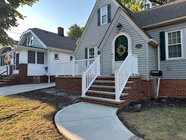 A grey house with a white railing porch, brick base, and a new concrete walkway leading to the front stairs.