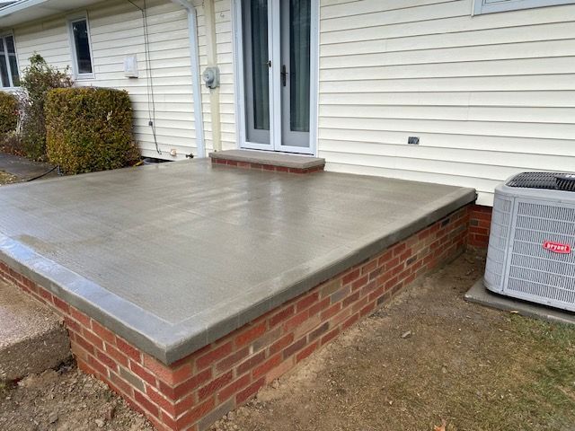 A new concrete patio with a brick foundation sits outside a house, located near an air conditioning unit.