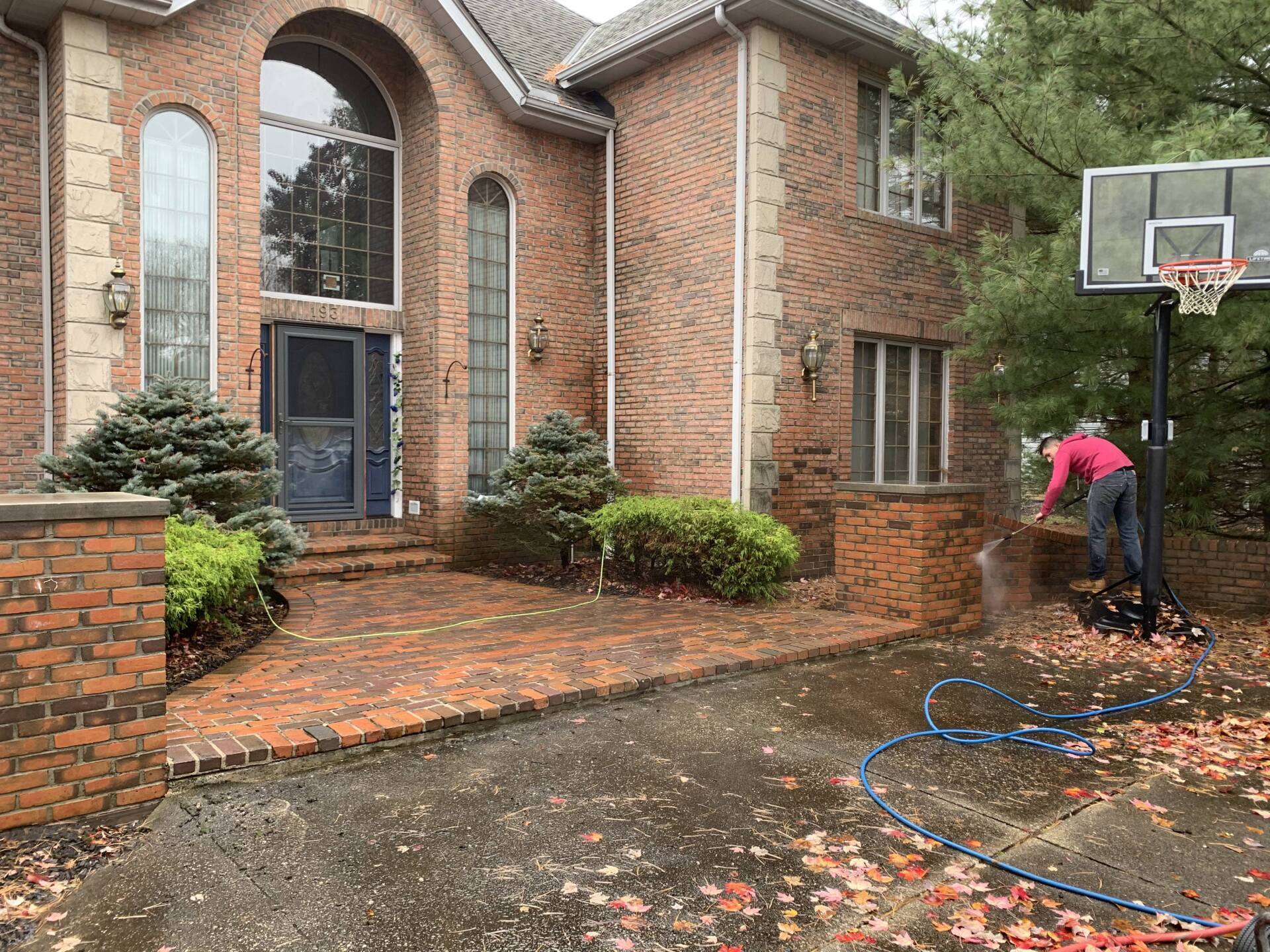 Person washing a brick driveway in front of a brick house with a basketball hoop. Fall leaves cover the ground.