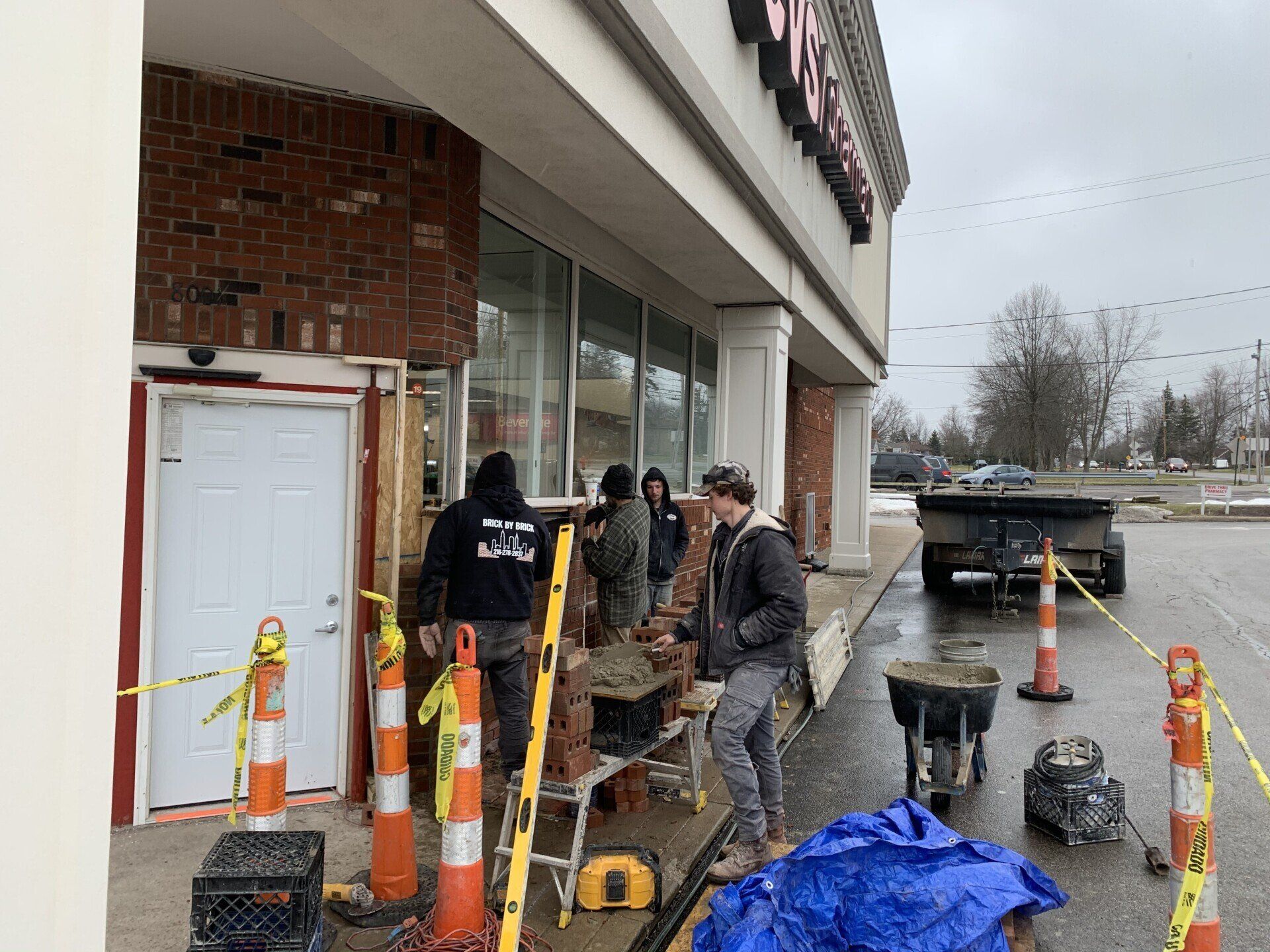 Construction workers repairing brickwork outside a store on a cloudy day. Orange cones and caution tape mark the work zone.